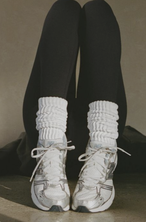 Legs raised against a plaster wall in black leggings, slouchy white ribbed pilates socks and white sneakers